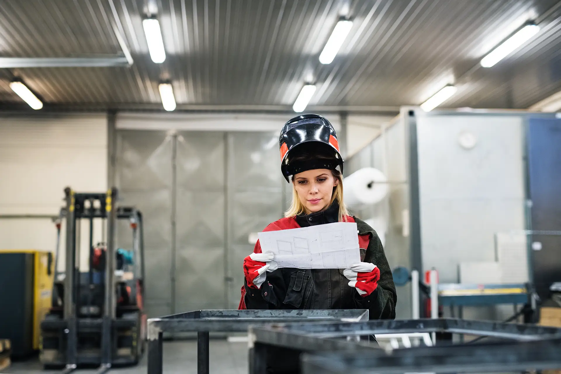Frau arbeitet als Schweißerin in einer Werkstatt, liest Baupläne und trägt Schutzkleidung und eine Schweißermaske.